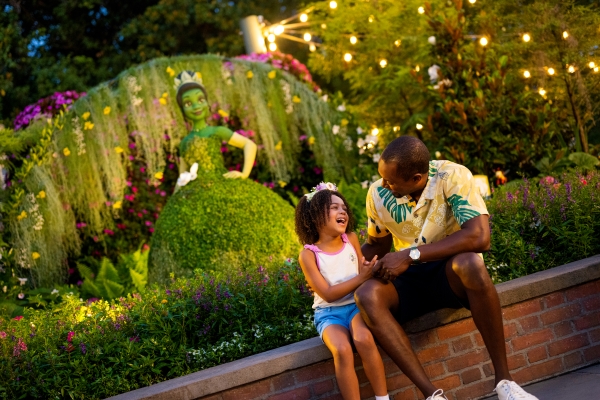 A dad and his daughter sit on a short brick wall together in front of a lit garden display featuring a topiary of Princess Tiana at EPCOT in the spring.