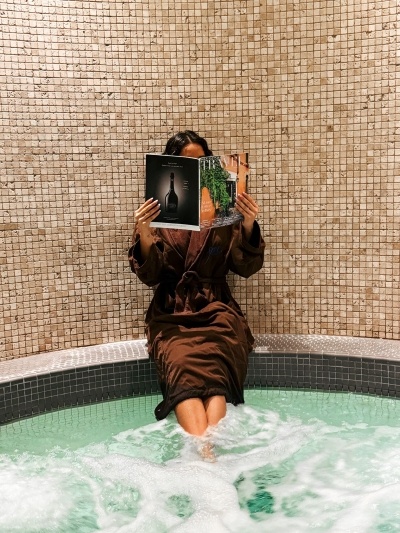 A woman in a brown silk robe reading a magazine with her feet in a whirlpool in front of a beige tiled wall. 