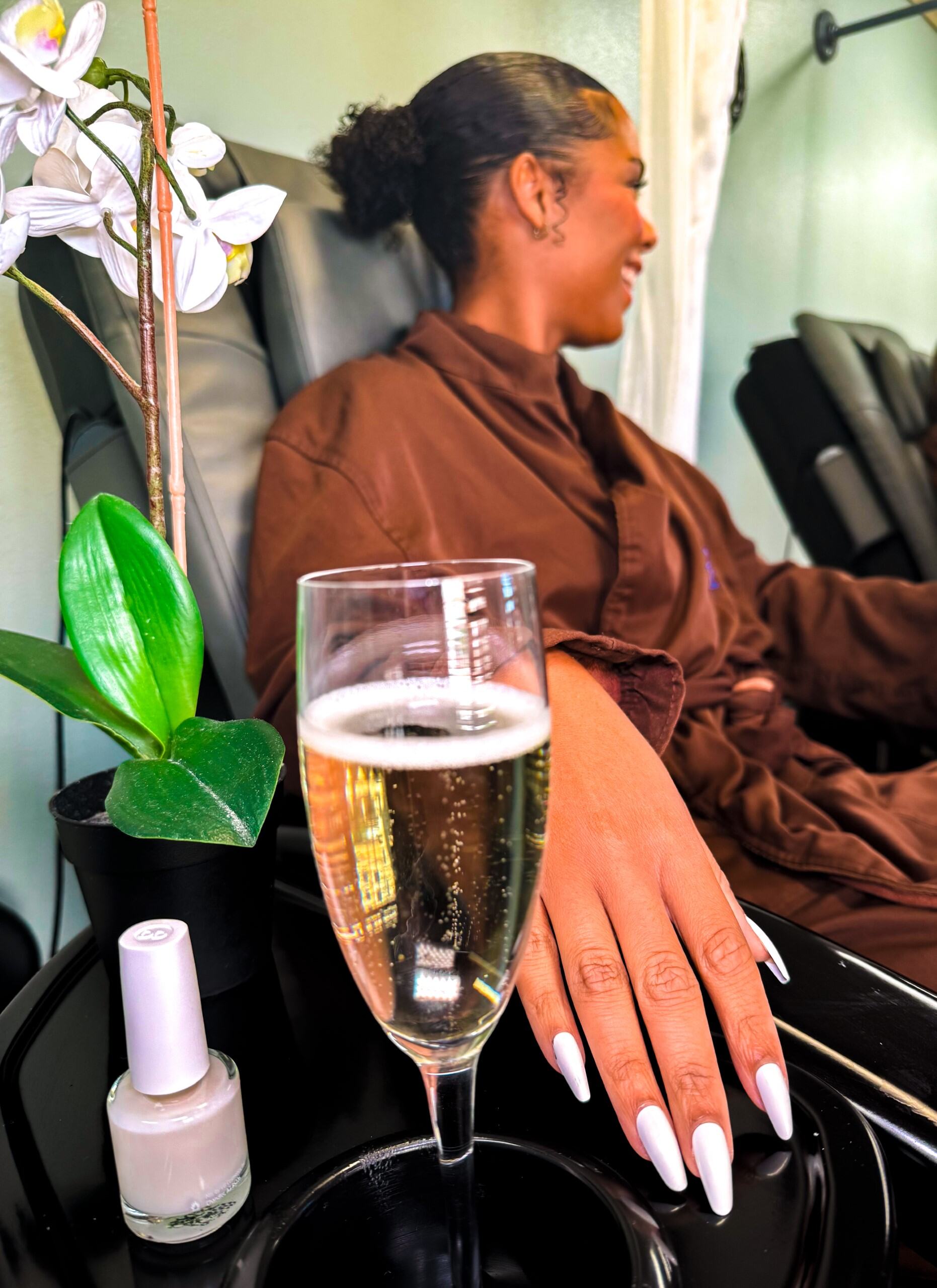 A Perfect Spa Day at Rosen Shingle Creek: What to Expect 6 A young woman in a brown spa robe looks away from the camera, sitting beside a table with a bottle of nail polish and a glass of Champagne where her manicured hand rests.
