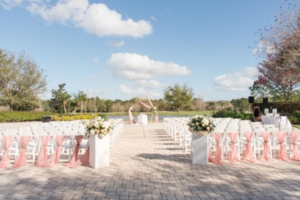 8 Questions to Ask When Choosing a Wedding Venue 8 A beautiful stone courtyard is set up with white chairs, pink floral arrangements, pink drapery, and a geometric archway draped in pink set against a backdrop of a golf course and serene pond.