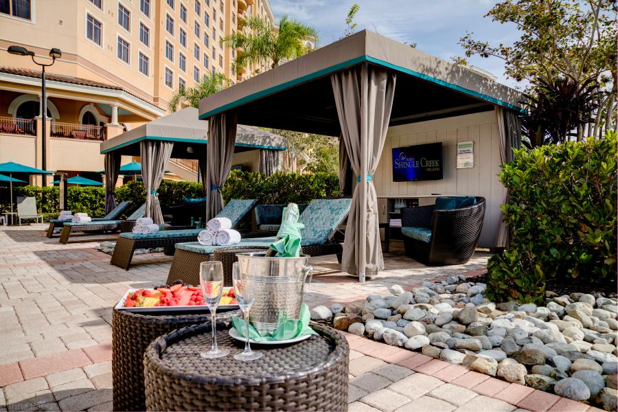 A shady cabana with curtains and blue cushioned lounge chairs, with a wicker table, Champagne, and fruit plate in the foreground. 