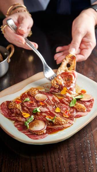 Hands holding a fork and a slice of bread, helping themselves to delicious carpaccio garnished with microgreens at Rosen Shingle Creek. 
