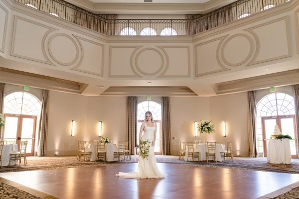 8 Questions to Ask When Choosing a Wedding Venue 7 A bride with a bouquet stands in the middle of beautiful two-story ballroom with Moulding, arched windows and doorways, and a dance floor.