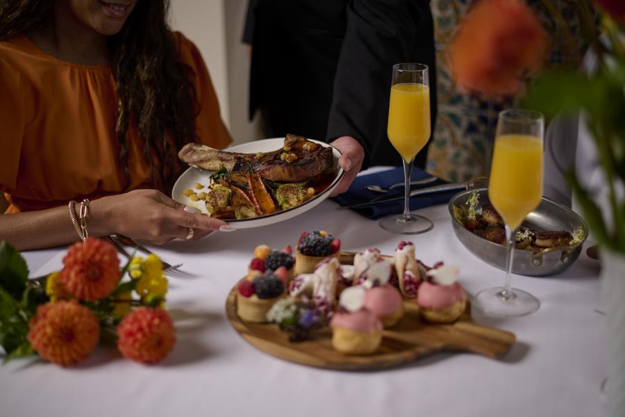 A waiter in black hands a plate with a large cut of meat and vegetables to a woman over a table with a mimosa, a wooden tray of fancy desserts, and a plate with sushi and charcuterie. 