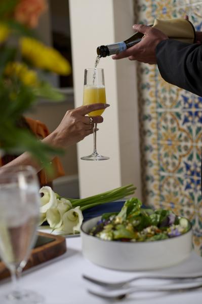 A woman’s hand holds a Champagne flute with a mimosa as a waiter pours Champagne over a table with a salad bowl and a bouquet of calla lilies. 