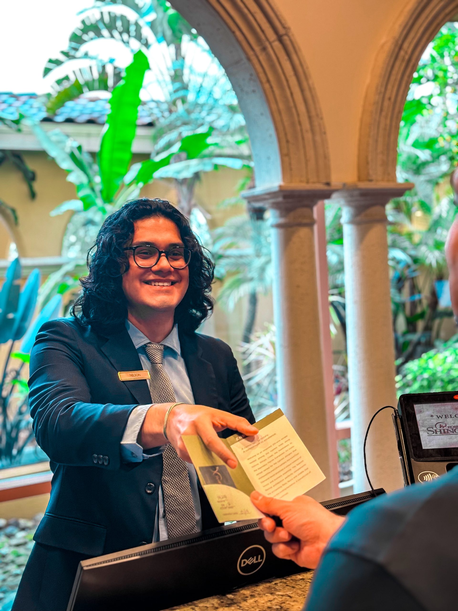 A man with long hair stands behind a front desk with arched windows, handing someone a room key.