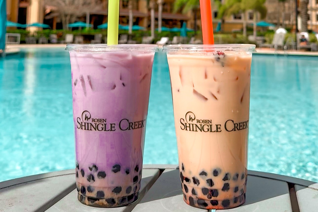 Two cups of boba tea, one purple and one tan in cups labeled Rosen Shingle Creek, sitting on a table beside a pool. 