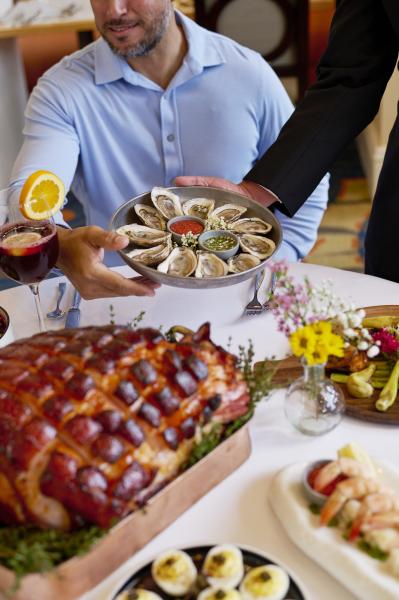 A waiter places a tray of oysters in front of a seated guest at a table with ham, yellow flowers, and other dishes.