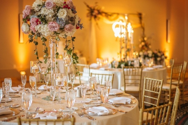 8 Questions to Ask When Choosing a Wedding Venue 2 An event set up with gold Chiavari chairs, ivory linens, glassware, tall floral centerpieces, and a neon light glowing out of focus in the background.