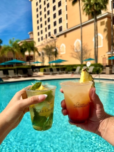 Two hands holding cocktails in cups that say "Rosen Shingle Creek" in front of a sparkling blue pool. 