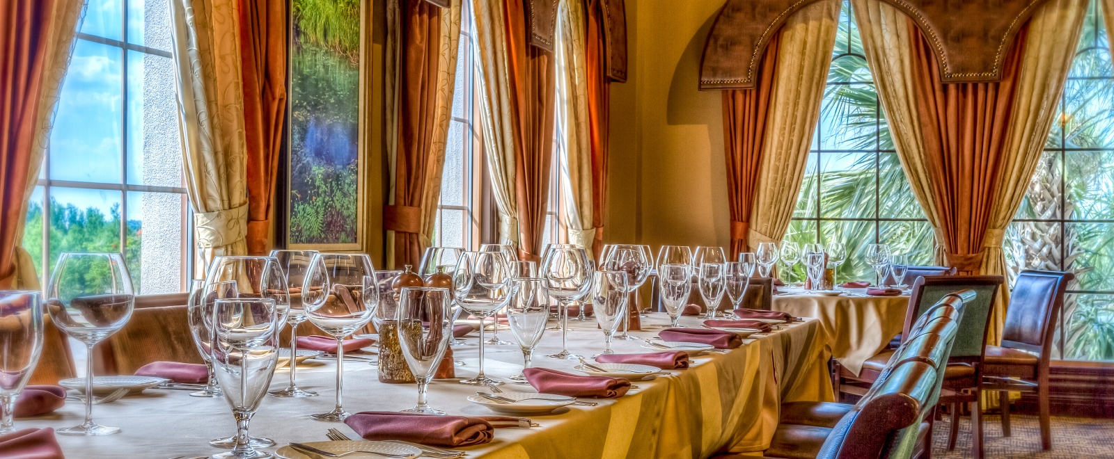 A beautifully set dining table with glassware in a room surrounded by windows overlooking tropical foliage with elegant curtains
