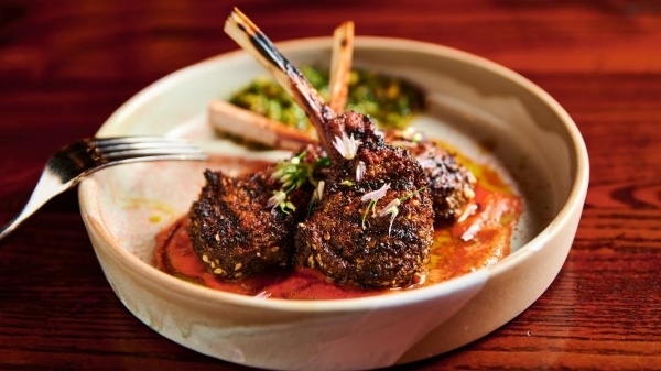 A round ceramic dish with stacked crusted lamb chop lollipops sitting on a red sauce with a fork resting on the side of the dish.