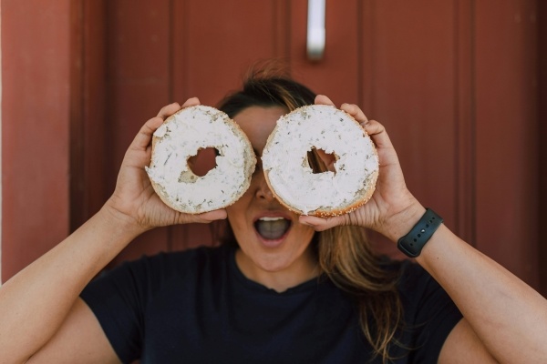 A woman in a black t-shirts holds two halves of a bagel with cream cheese in front of her face with a surprised expression.