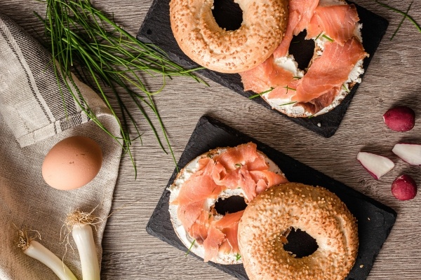 An above shot of bagels with cream cheese and lox sitting on a wooden surface beside chives, an egg, and green onions. 