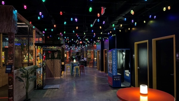 A dimly lit room with colored lights hanging on the ceiling, a photo booth, high top tables, and caged spaces for axe throwing.