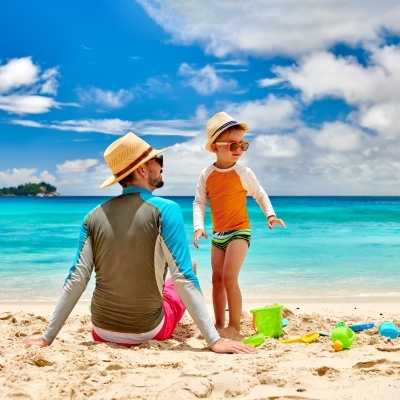 A man and his son, both wearing long sleeve rash guards, sit on the beach in front of clear blue water.