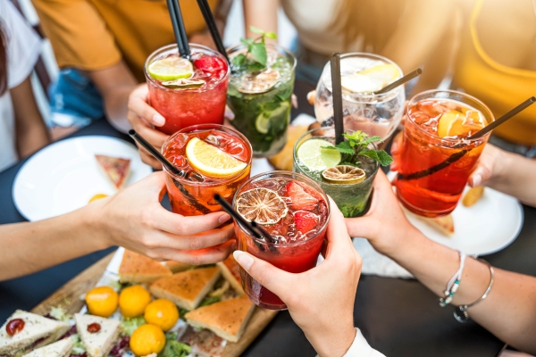 An overhead shot of hands toasting with colorful cocktails over a table set with delicious plates of food. 