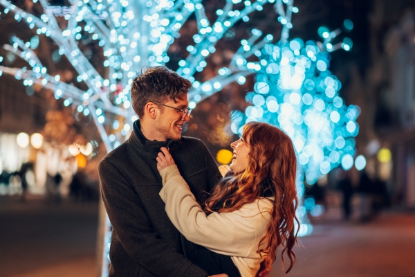 A couple in jackets laugh and embrace in front of a tree full of Christmas lights while enjoying a romantic holiday date night in Orlando.