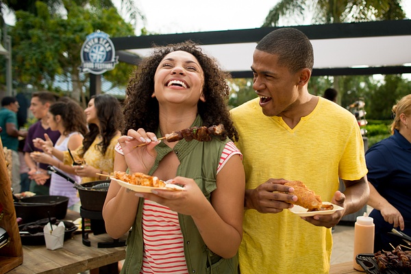 A laughing man and woman in a crowd holding small plates of food outdoors. 