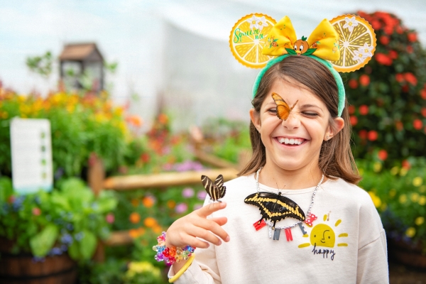 A young girl wearing citrus Minnie Mouse ears stands in a garden smiling, with three colorful butterflies resting on her finger, chest, and nose.