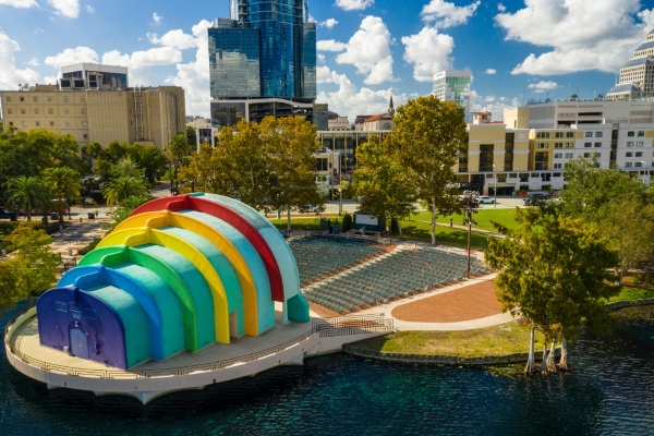 A rainbow-colored amphitheater hovering over a lake and surrounded by trees and tall buildings.
