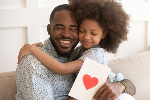 A young girl hugs her dad, who holds a card with a heart on the front.