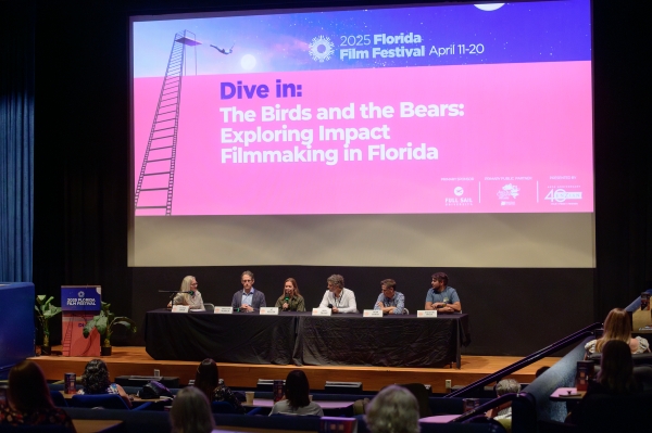 A panel of six people sitting at a long table on a stage in front of a  movie theatre screen. 