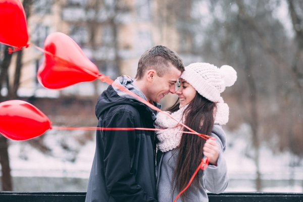 A couple wearing winter outfits marveling over each other while holding heart-shaped balloons.