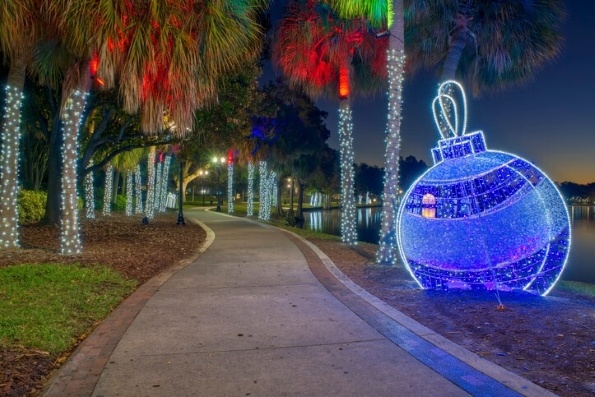 A pathway at night lined in palm trees decorated in white holiday lights and a large Christmas ornament decorated in lights. 