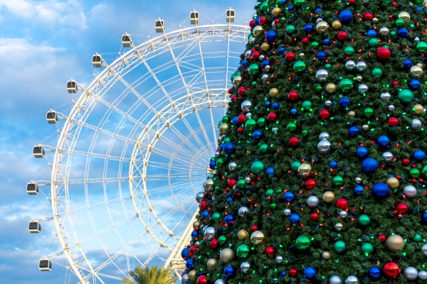 A Christmas tree in the foreground with a large ferris wheel against a blue cloudy sky behind it, representing the holiday evets at ICON Park in Orlando.