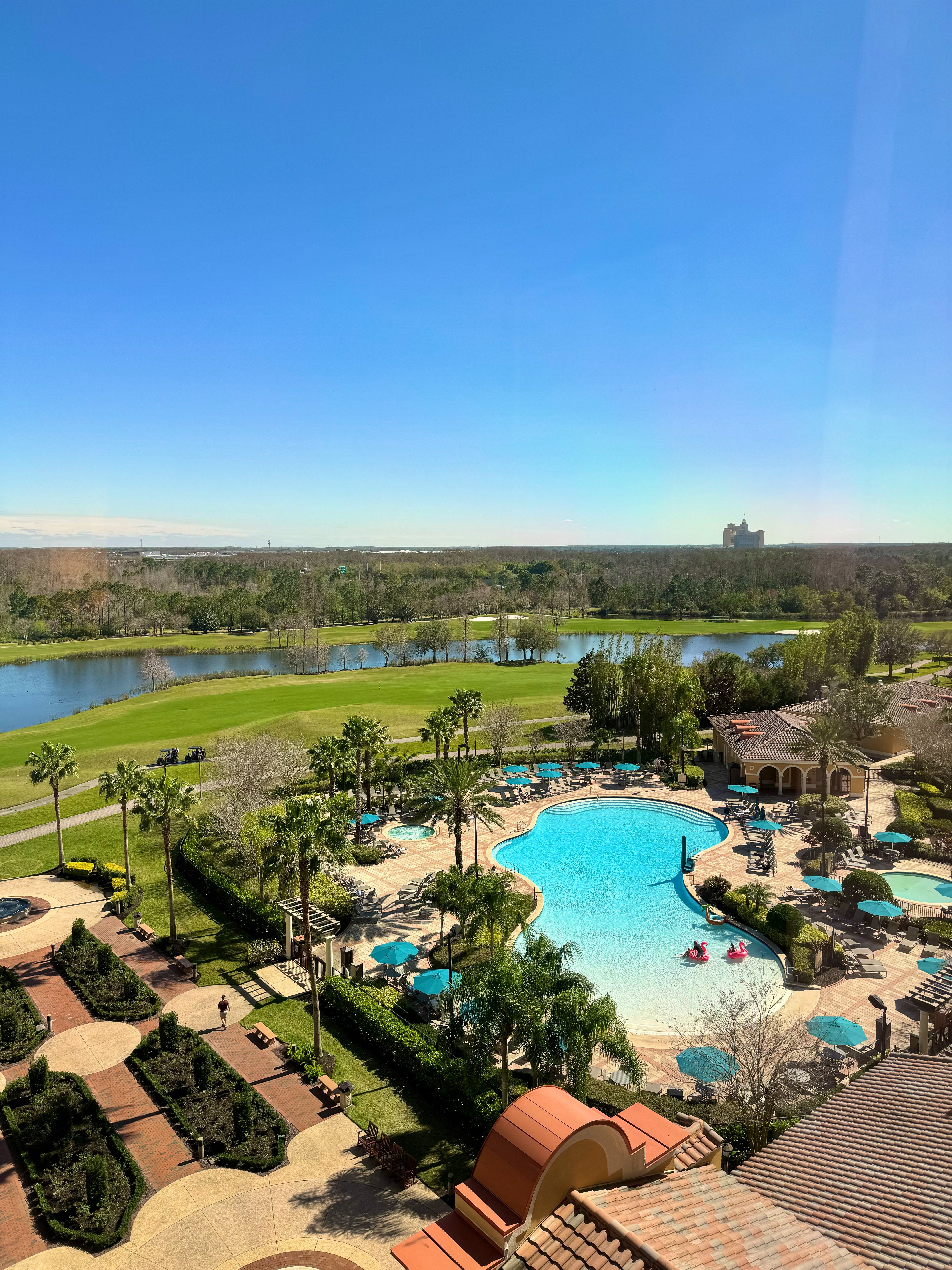  An elevated view of the Rosen Shingle Creek resort, showing a large pool surrounded by palm trees, lounge chairs with blue umbrellas, and a golf course with a lake in the background.