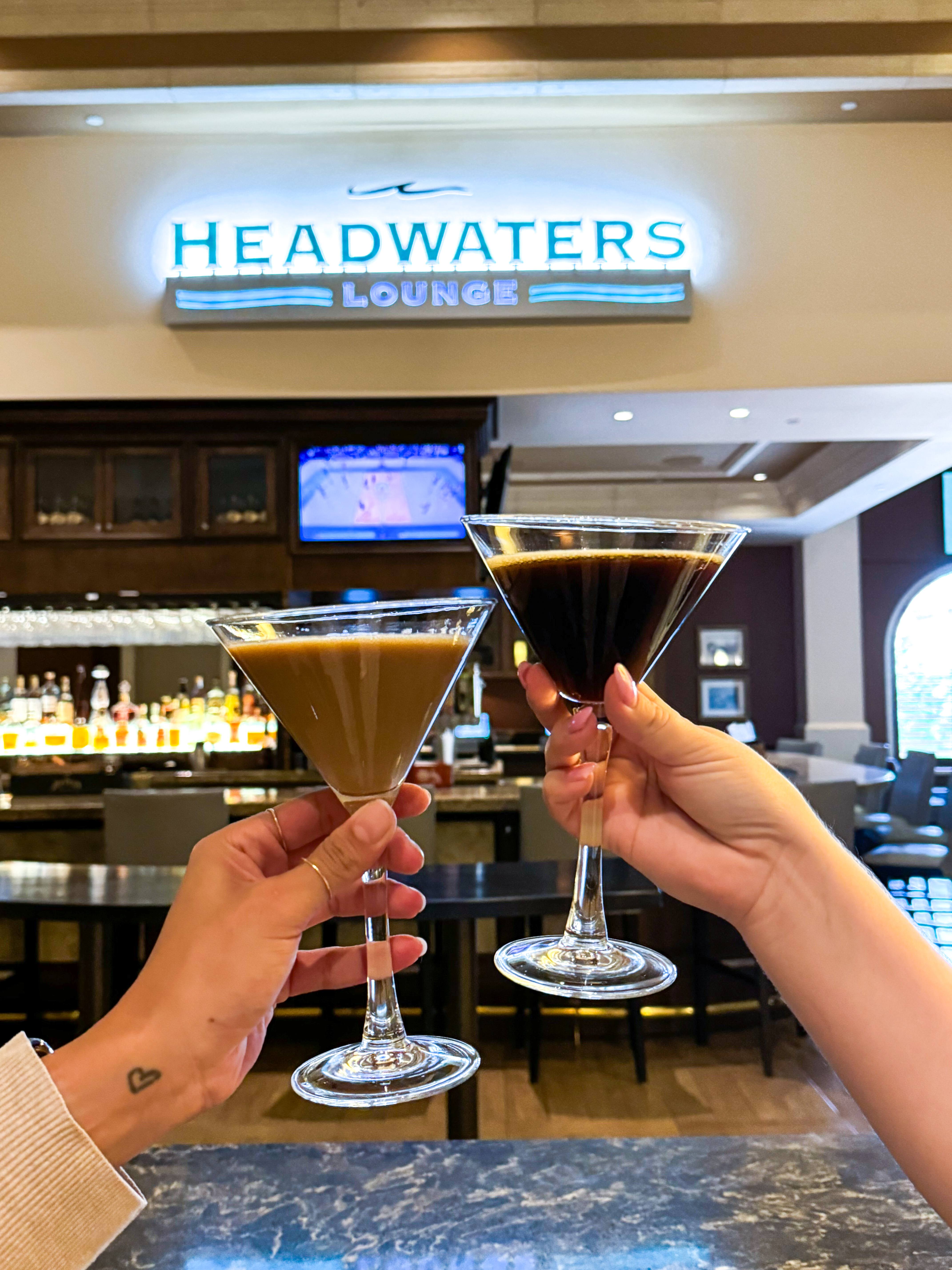 Two hands holding martini glasses filled with cocktails, one light brown and one dark, in front of the "Headwaters Lounge" sign at Rosen Shingle Creek -- a great location to explore International Drive's Premier Happy Hour Hotspots.