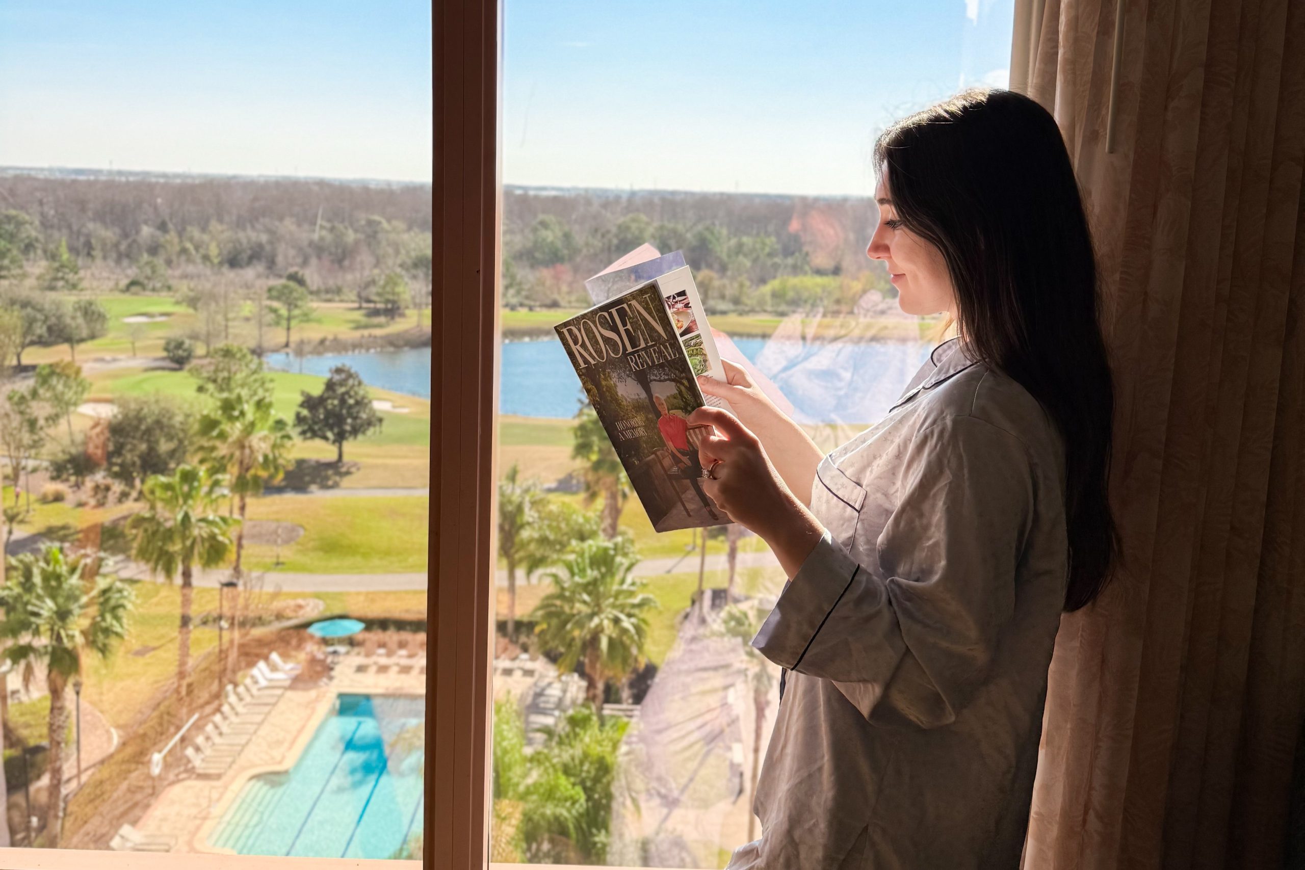 A woman reading a Rosen Reveal magazine in a hotel room leans beside a window overlooking a pool and a lake. 