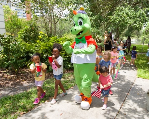 A green dinosaur mascot leads a group of children along a sidewalk in a park. 