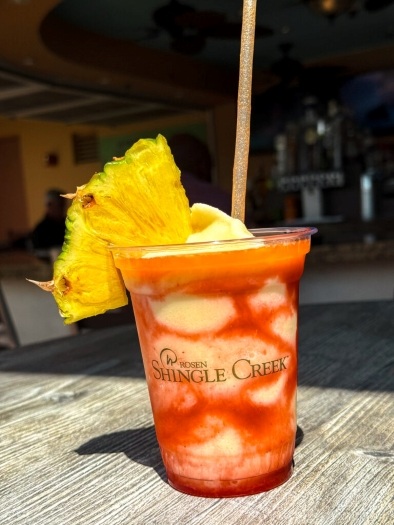 What Your Cocktail Order from Cat-Tails Pool Bar & Grille Says About You 4 A frozen white and red marbled cocktail with a straw and a large pineapple garnish in a plastic cup labeled Rosen Shingle Creek.