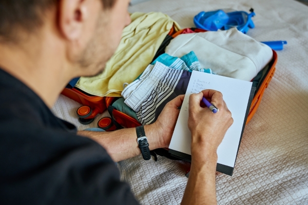 A man checks off a list while sitting beside a packed suitcase on a bed.