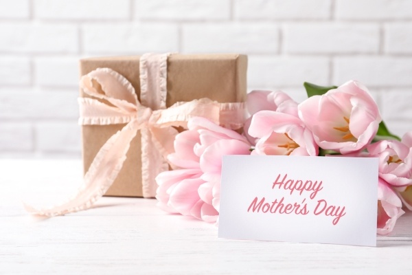 A bouquet of pink tulips beside a gift wrapped in brown parchment and a pink ribbon with a card that says ”Happy Mother’s Day.”