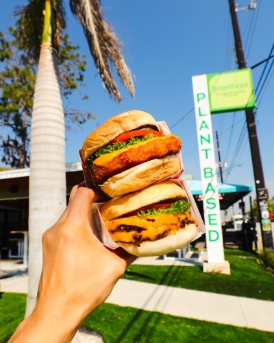 A hand holding a stacked burger and a chicken sandwich in front of Plantees, a vegan dining establishment in Orlando. 