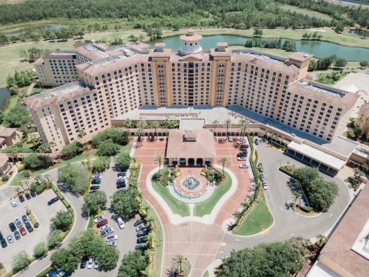 An aerial shot of a large hotel surrounded by a large pond, trees, and a beautiful golf course.