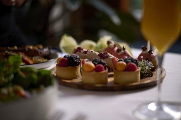 A selection of berry tarts and other patisserie on a round wooden tray with an out of focus salad bowl and mimosa in the foreground.