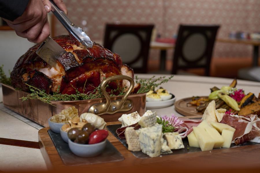 Hands carve a roast ham on a table behind a wooden charcuterie board displaying an array of meats, cheeses, olives, and vegetables.