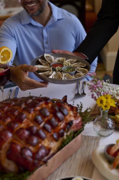 A man in a blue shirt takes a plate of oysters from a waiter in a black jacket, with a small vase of yellow and pink flowers and a large roast ham sitting on the table in front of him.