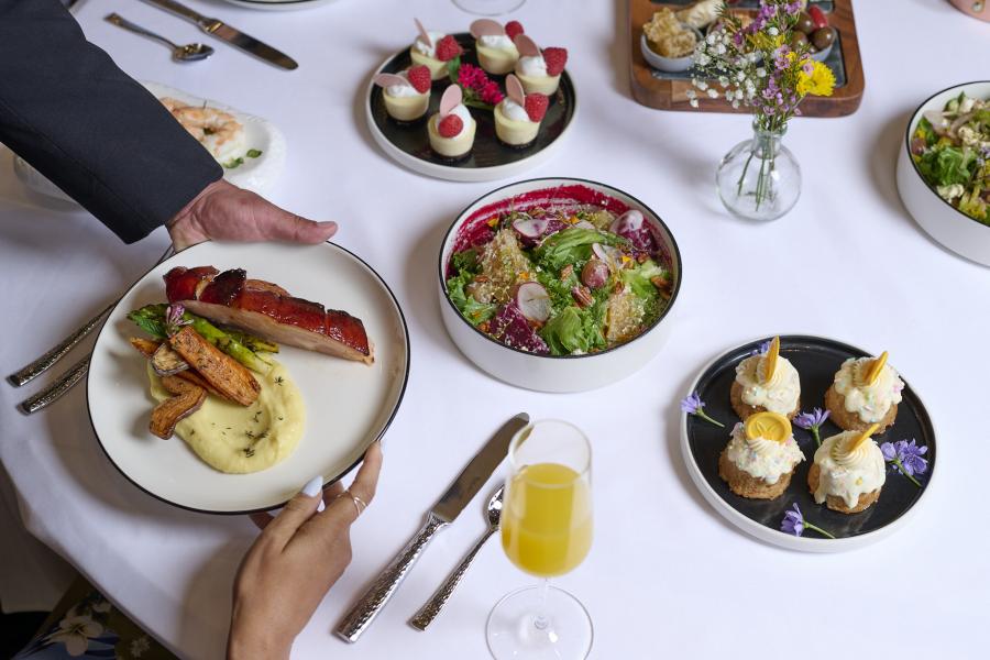 A hand in a black jacket passes a plate of food to a woman at a table with a mimosa, salad bowls, and two plates of elegant tarts. 