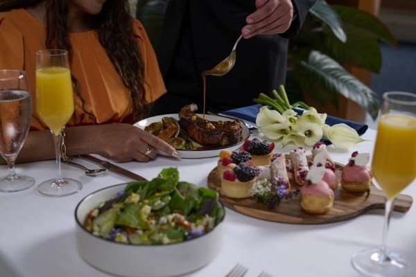 A woman in an orange top holds a plate with a large steak as a waiter drizzles sauce on top. The table is set with mimosas, a bowl of salad, and a round wooden tray of desserts. 
