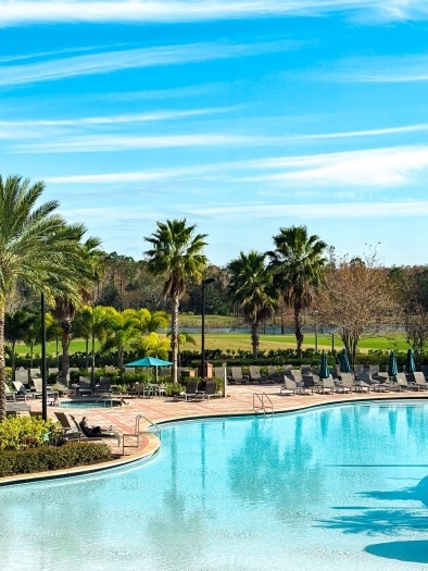 A large sparkling blue pool surrounded by lounge chairs and palm trees, with a golf course in the distance. 