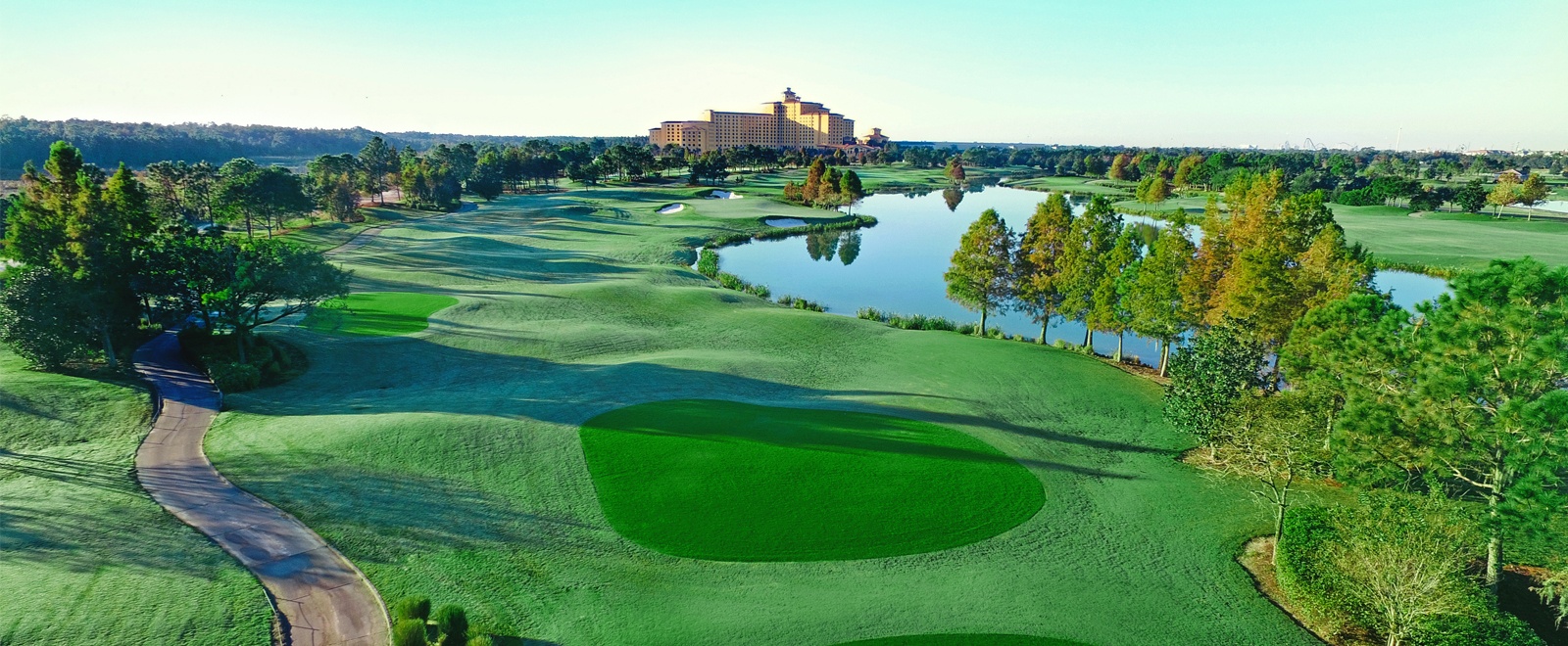 An aerial view of a sprawling golf course with green lawns, mature trees, and a lake with a hotel in the distance