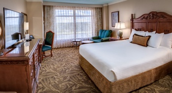 A bright hotel room with a large bed with a wooden headboard, windows overlooking a golf course, and a blue sofa