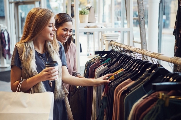 Two women browse through a rack of clothing at a store with cups of coffee and shopping bags.