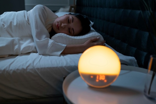 A woman sleeps in a bed at night next to a nightstand with a glowing alarm clock night light.
