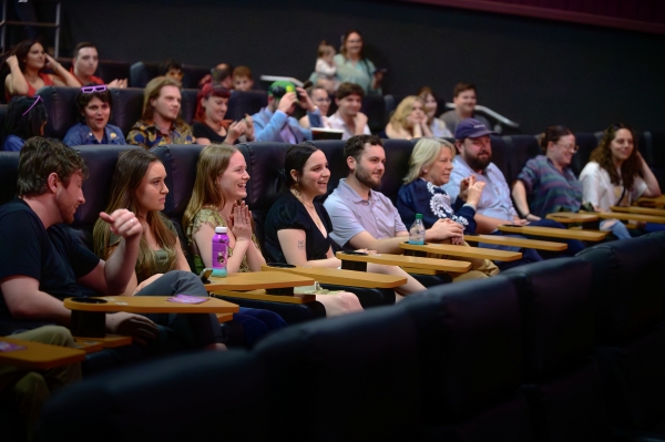 A row of smiling people sitting in movie theatre seats with tables. 